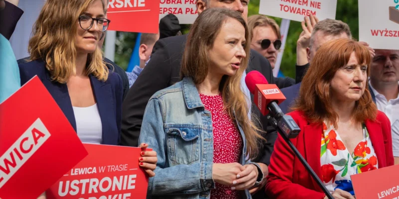 Grupa osób na demonstracji politycznej trzyma czerwone plakaty Lewica; na pierwszym planie kobieta w jeansowej kurtce wygłasza przemówienie do mikrofonu, a obok niej stoi kobieta w czerwonej marynarce.