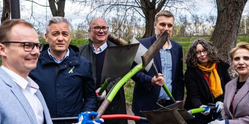 Na zdjęciu grupa osób pozująca z łopatami w parku w słoneczny dzień.