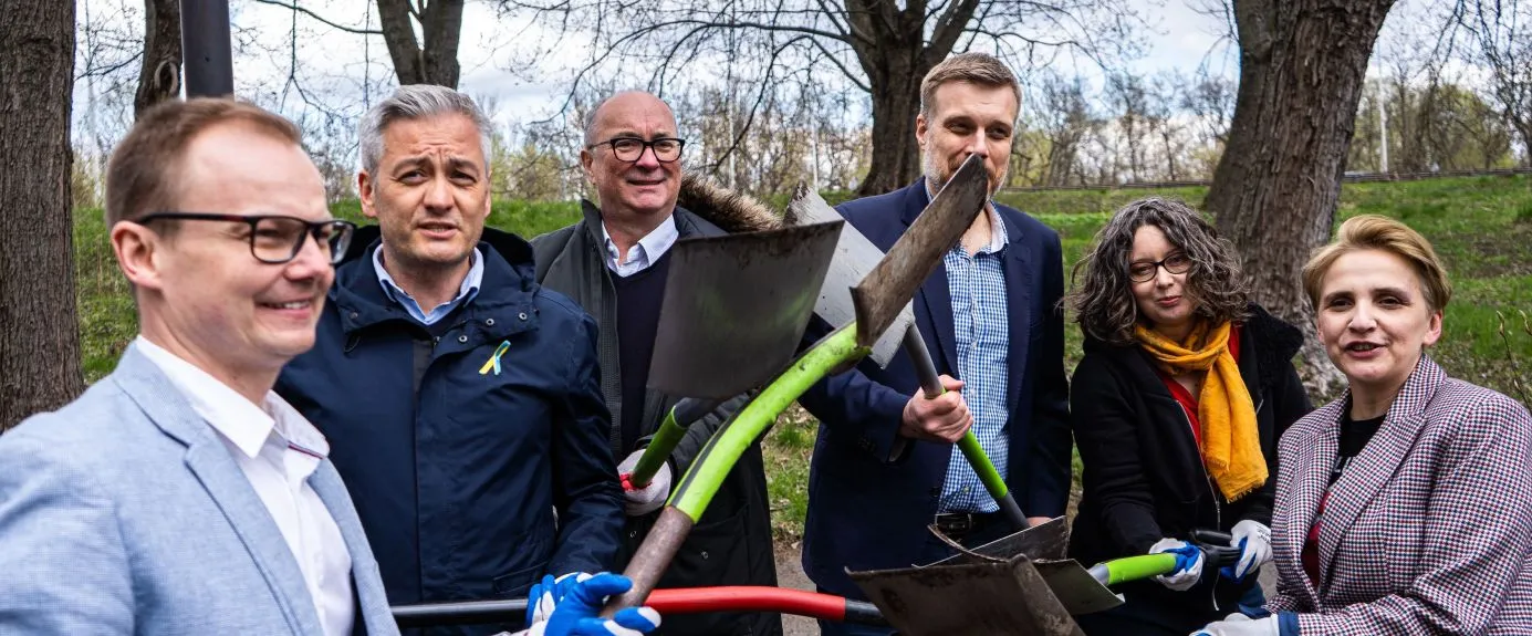 Na zdjęciu grupa osób pozująca z łopatami w parku w słoneczny dzień.