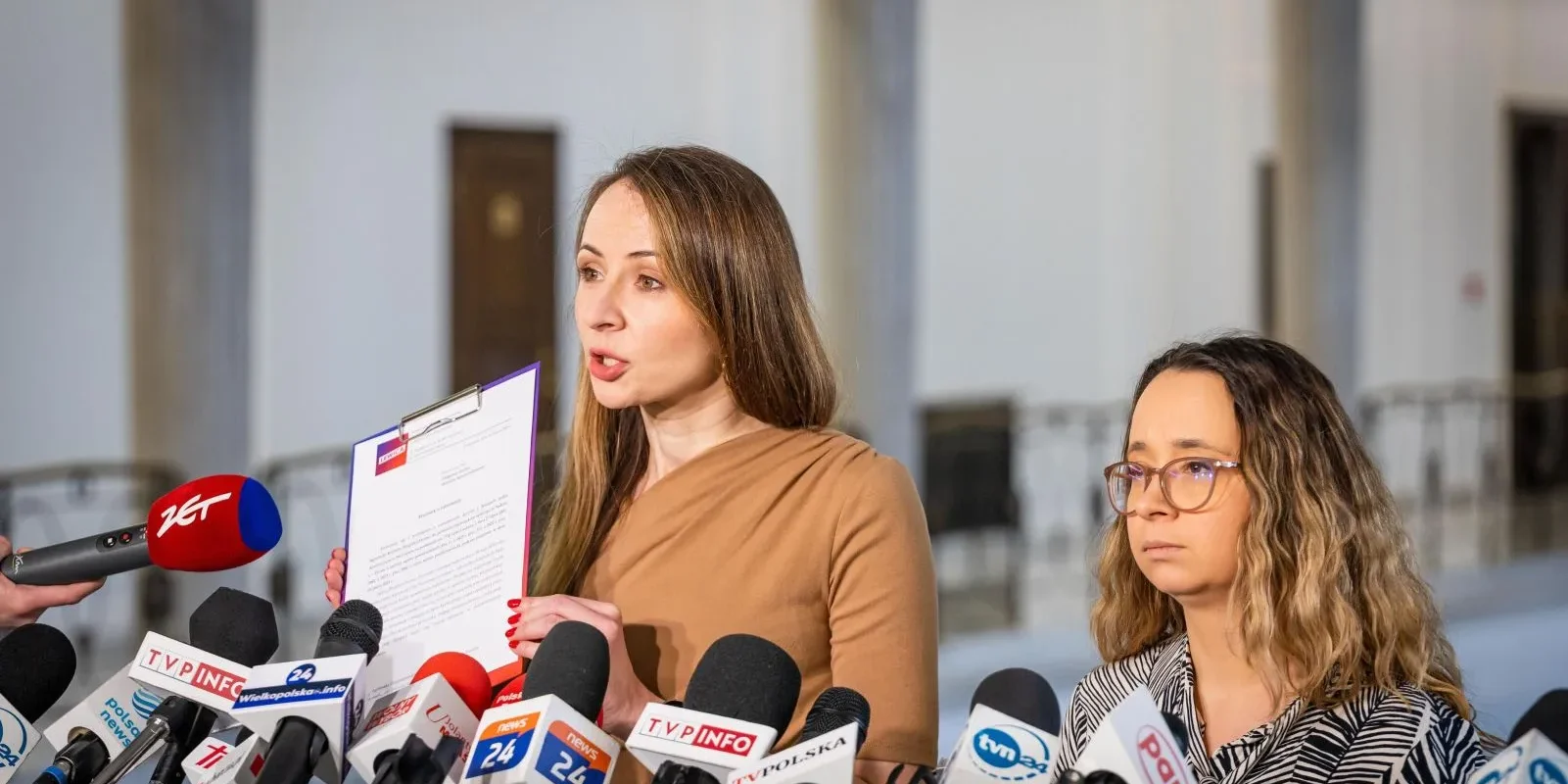 Na zdjęciu widać kobietę mówiącą do kamer, trzymającą dokument, obok niej stoi druga kobieta, otoczone mikrofonami i kamerami w tle.