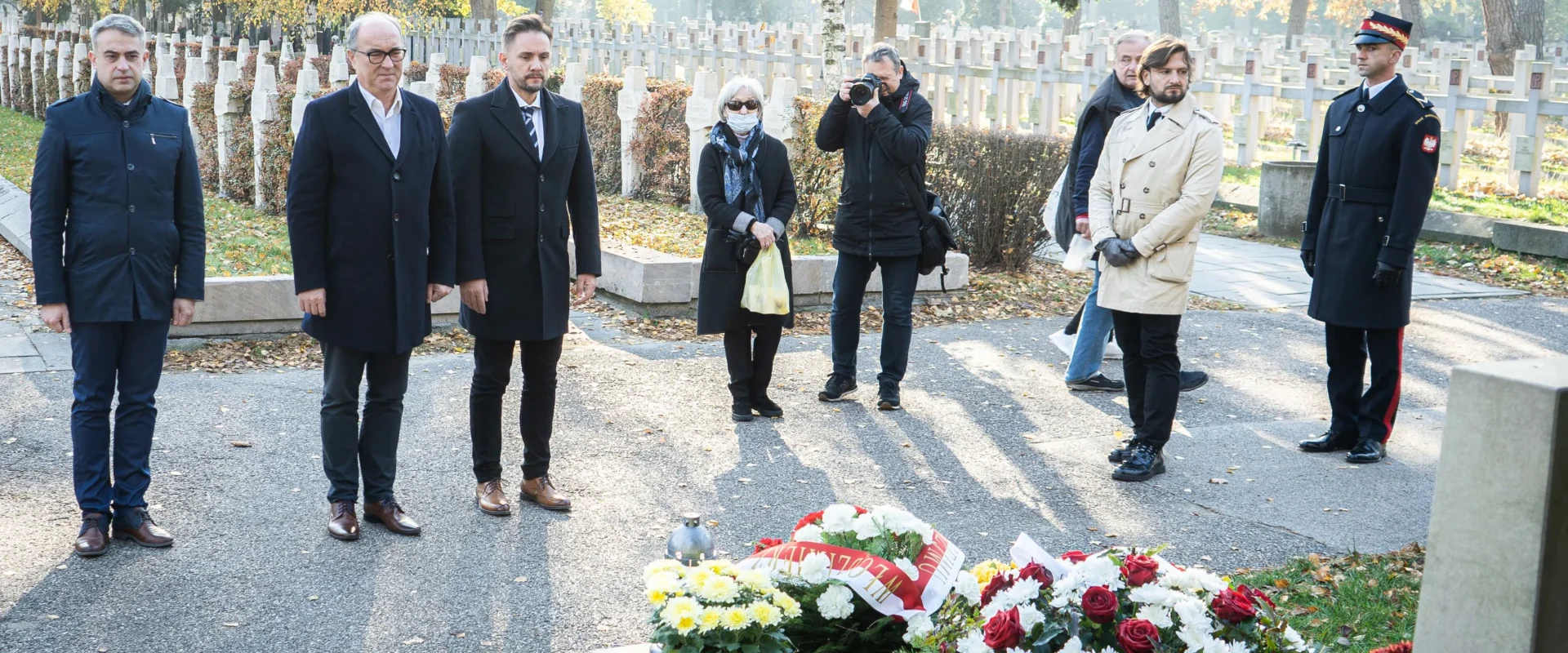 Grupa osób w eleganckich ubraniach stoi w milczeniu przy grobie, obok którego znajdują się kwiaty i znicze, w otoczeniu jesiennego krajobrazu.