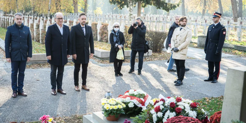 Grupa osób w eleganckich ubraniach stoi w milczeniu przy grobie, obok którego znajdują się kwiaty i znicze, w otoczeniu jesiennego krajobrazu.