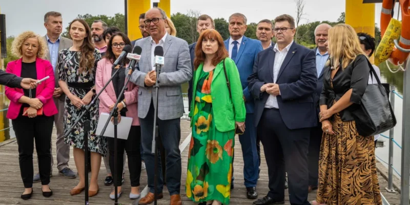 Grupa ludzi w formalnych ubraniach stoi na tle wody, otoczona żółtymi elementami infrastruktury, podczas konferencji prasowej.