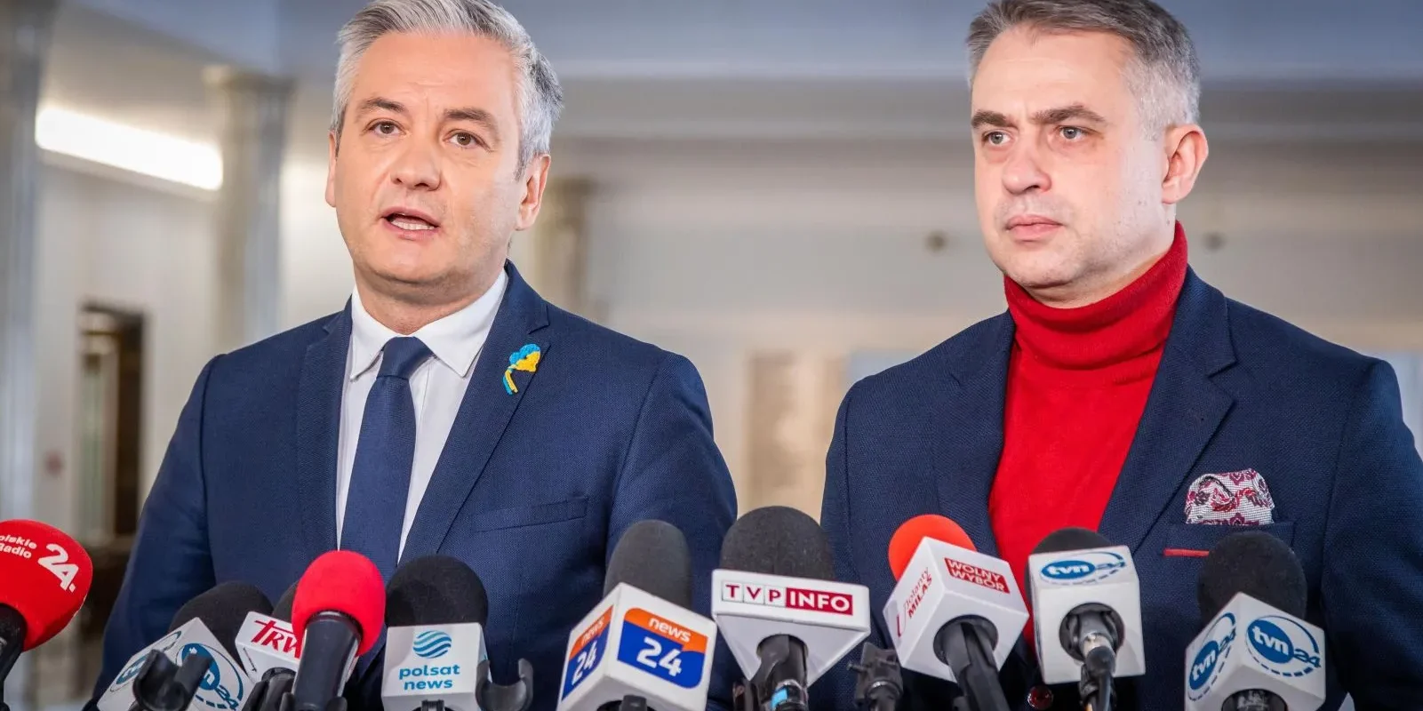 Dwóch mężczyzn w formalnych strojach stoi przed mikrofonami podczas konferencji prasowej w jasnym wnętrzu.