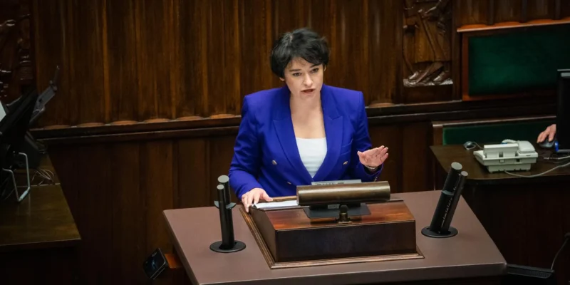 Na zdjęciu widoczna jest kobieta w niebieskim żakiecie, mówiąca z mównicy w sali obrad, z drewnianą ścianą w tle.