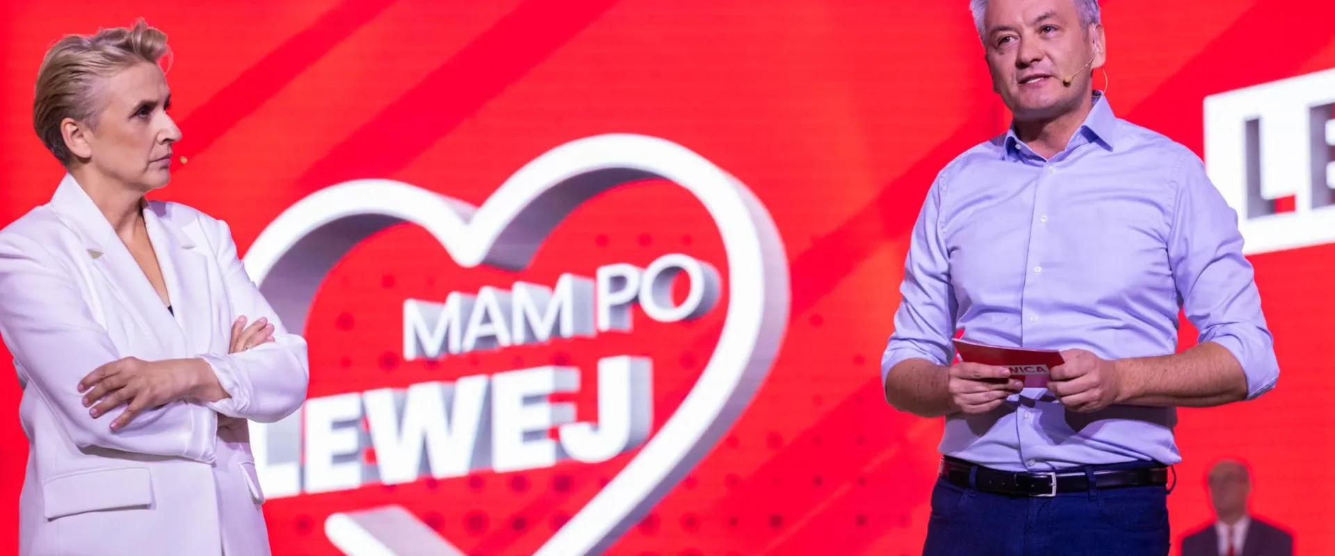 Dwóch prowadzących na scenie przed czerwonym tłem z sercem i napisem, kobieta w białej marynarce po lewej, mężczyzna w niebieskiej koszuli z mikrofonem po prawej, omawiają projekt związany z 35-godzinnym tygodniem pracy.