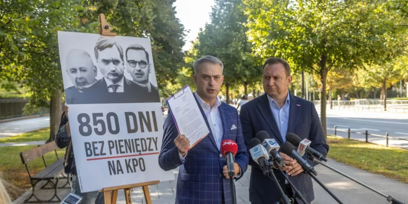 Dwóch mężczyzn w garniturach udziela konferencji prasowej w parku, stojąc za mikrofonami, a obok nich znajduje się plakat z trzema mężczyznami i napisem „850 DNI BEZ PIENIEDZY NA KPO”.