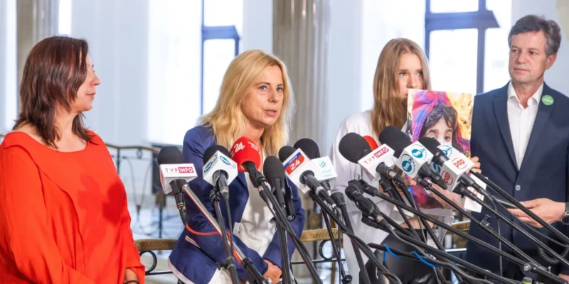 Na zdjęciu widać grupę osób podczas konferencji prasowej, z mikrofonami w tle i jedną osobę trzymającą zdjęcie dziecka.