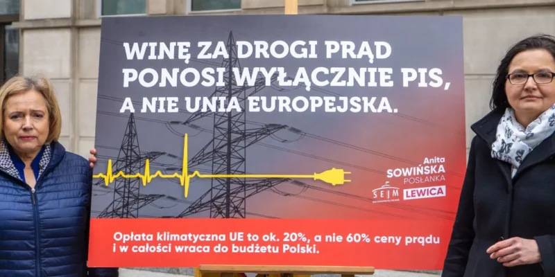 Dwie kobiety stoją po obu stronach dużego protestacyjnego plakatu o wysokich kosztach prądu, z tłem w postaci linii energetycznych i słupów napowietrznych.