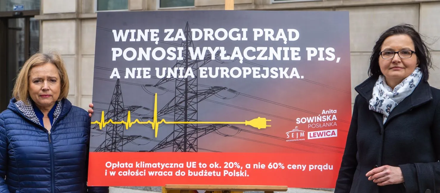 Dwie kobiety stoją po obu stronach dużego protestacyjnego plakatu o wysokich kosztach prądu, z tłem w postaci linii energetycznych i słupów napowietrznych.
