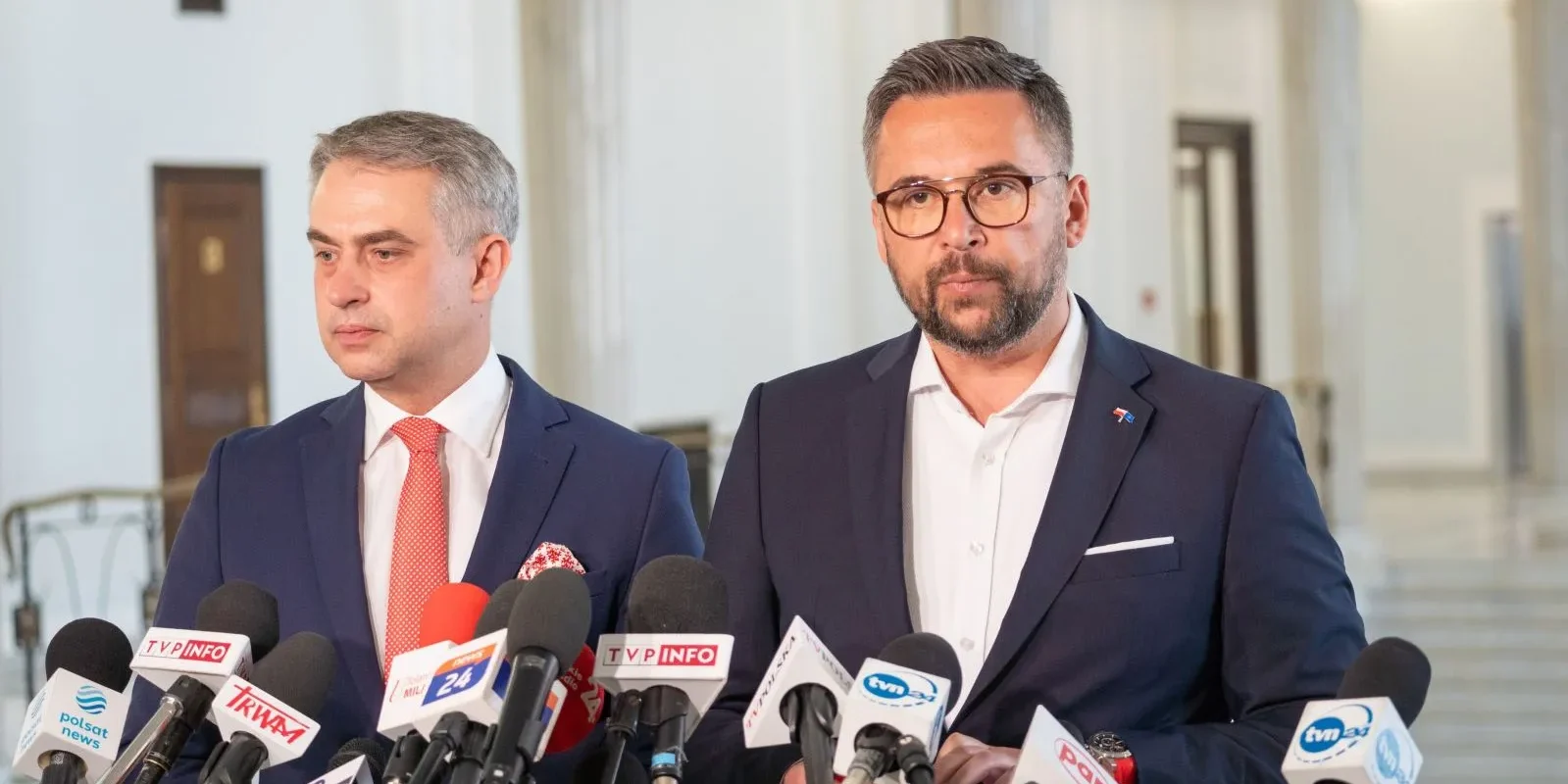 Dwóch mężczyzn w garniturach podczas konferencji prasowej, stojących przy licznych mikrofonach dziennikarzy w holu budynku.