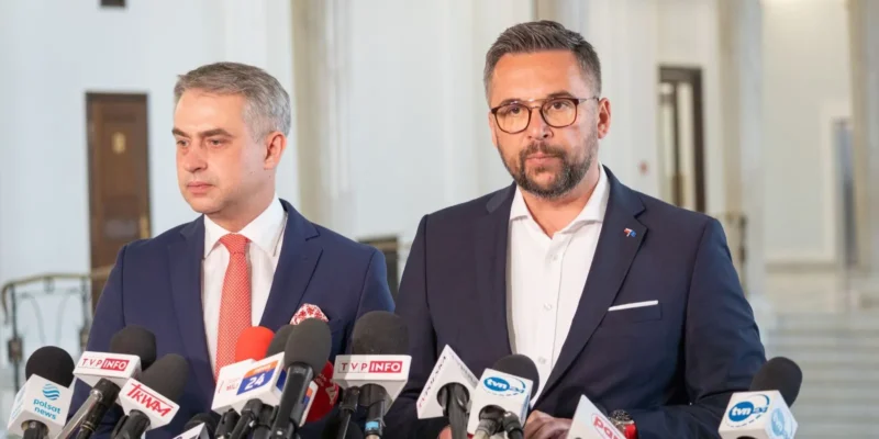 Dwóch mężczyzn w garniturach podczas konferencji prasowej, stojących przy licznych mikrofonach dziennikarzy w holu budynku.