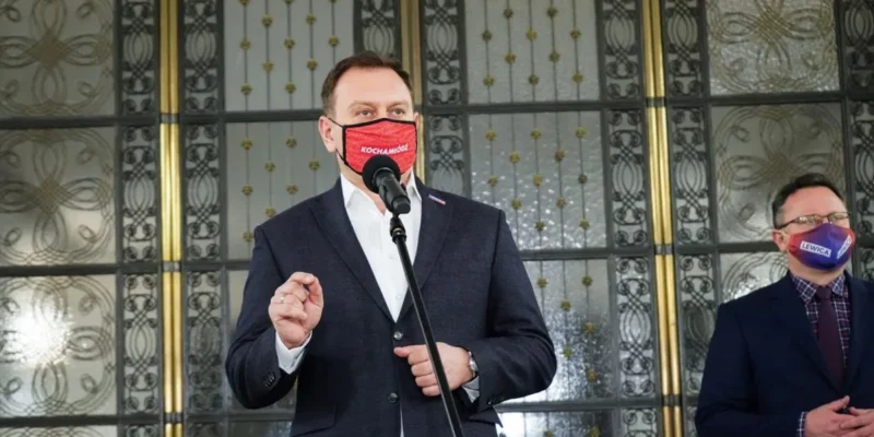 Dwoje mężczyzn w garniturach stoi przy mikrofonie, jeden przemawia, oboje noszą maseczki z napisami, w tle ozdobne drzwi.