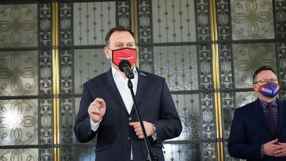 Dwoje mężczyzn w garniturach stoi przy mikrofonie, jeden przemawia, oboje noszą maseczki z napisami, w tle ozdobne drzwi.