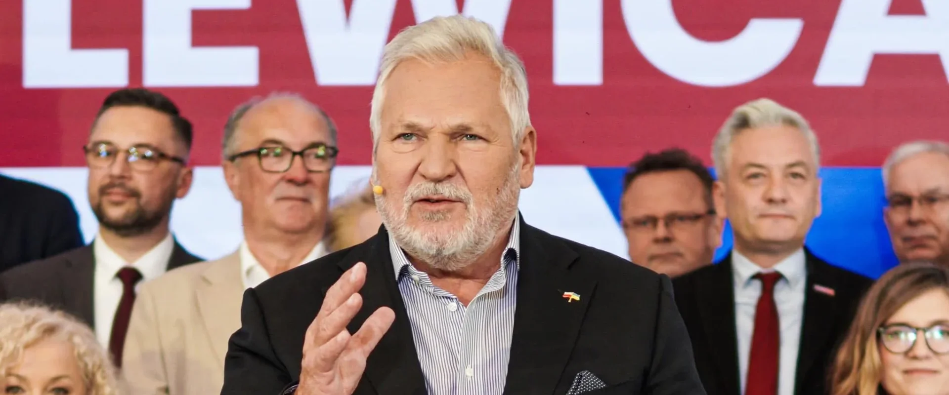 Mężczyzna w czarnym garniturze przemawia na scenie, na tle dużego banera z napisem Lewica i stojących za nim osób.