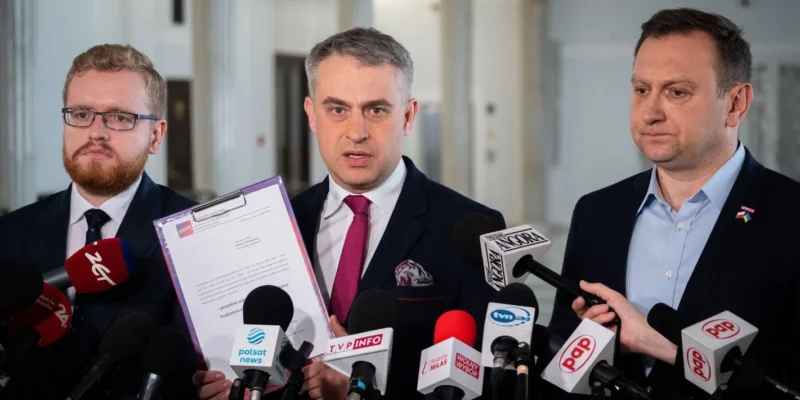 Trzech mężczyzn w garniturach podczas konferencji prasowej; w centrum mężczyzna prezentuje dokument w fioletowej teczce, otoczony mikrofonami mediów.