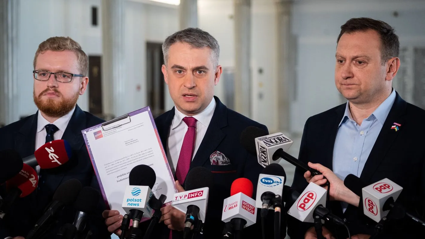 Trzech mężczyzn w garniturach podczas konferencji prasowej; w centrum mężczyzna prezentuje dokument w fioletowej teczce, otoczony mikrofonami mediów.