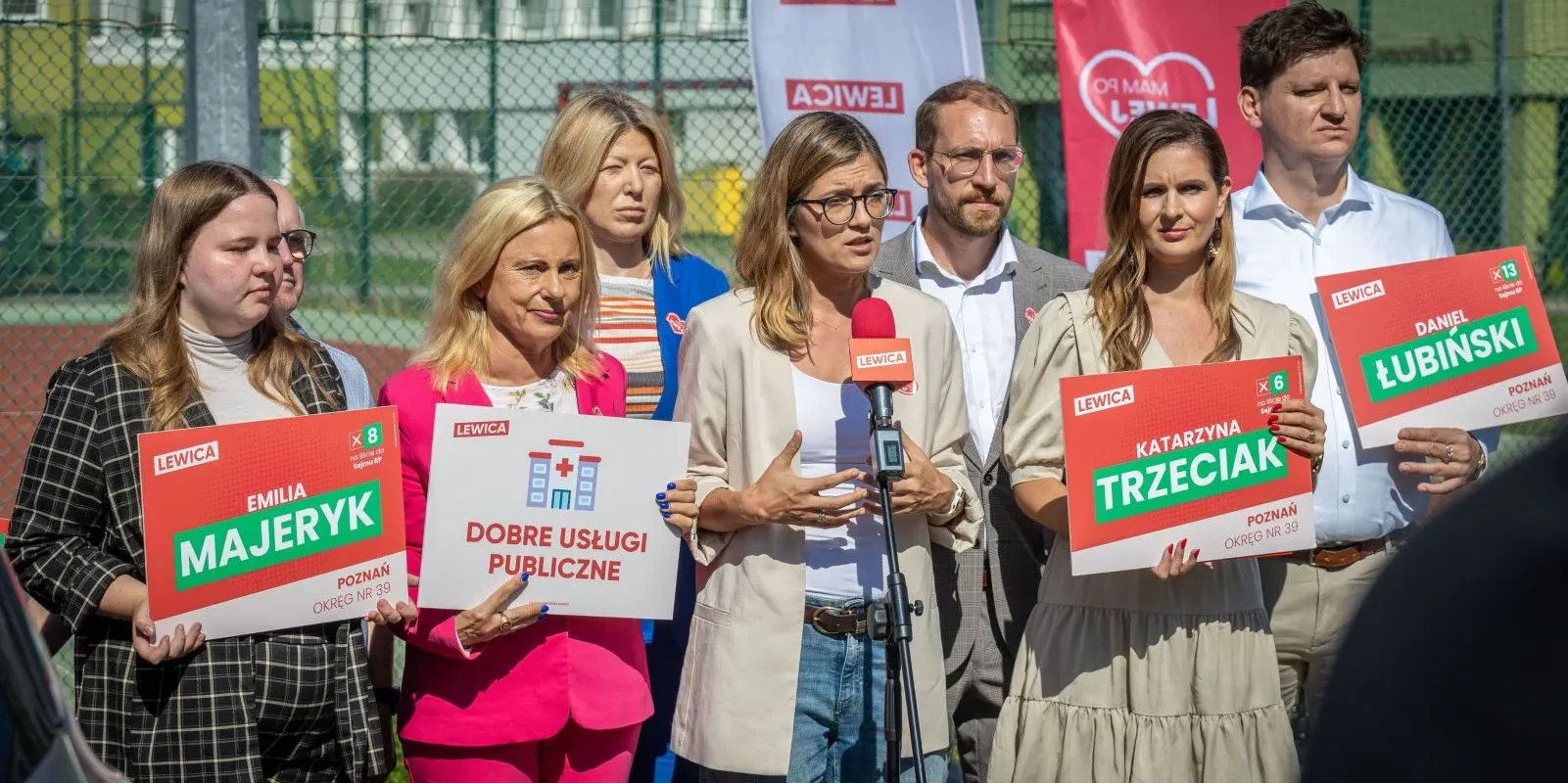 Grupa osób na zewnątrz przy ogrodzeniu, trzymających plakaty wyborcze Lewicy z nazwiskami kandydatów i hasłami, podczas gdy jedna kobieta przemawia do mikrofonu.