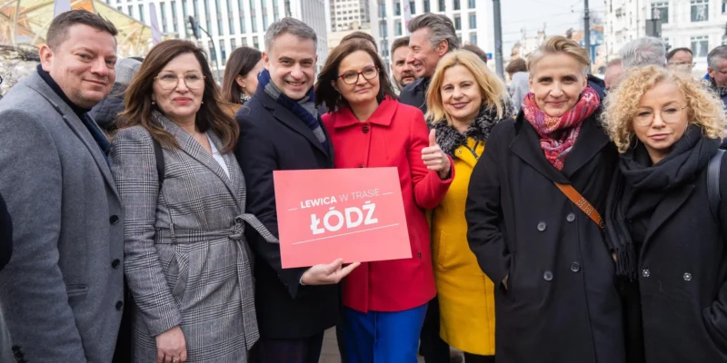 Grupa osób w zimowych ubraniach stoi na placu w Łodzi, trzymając czerwony plakat z napisem „LEWICA W TRASIE ŁÓDŹ”.