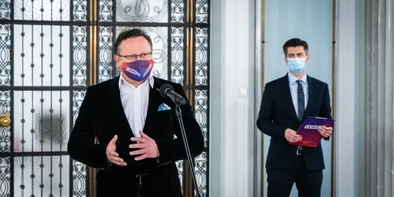 Dwóch mężczyzn w garniturach stoi w eleganckim wnętrzu z ozdobnymi przęsłami; jeden po lewej przemawia do mikrofonu, ma czarną marynarkę i gradientową maseczkę z napisem LEWICA, a drugi po prawej trzyma fioletową broszurę z tym samym napisem.