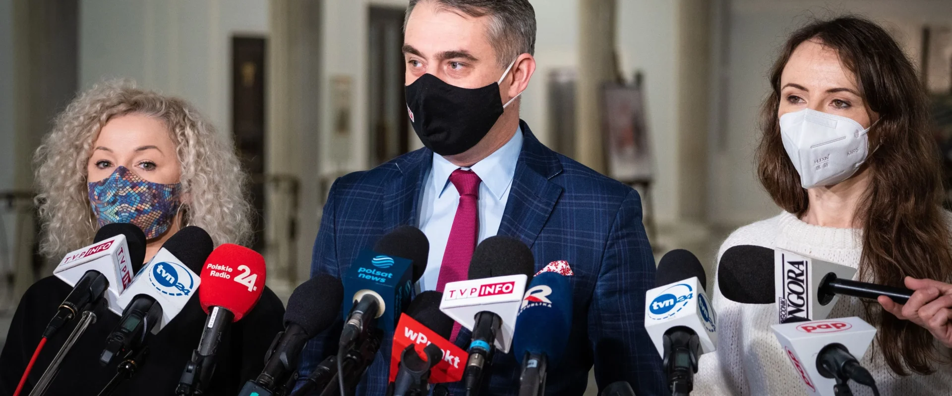 Dwie kobiety i jeden mężczyzna w maseczkach udzielają konferencji prasowej Lewicy, stojąc za zestawem mikrofonów.