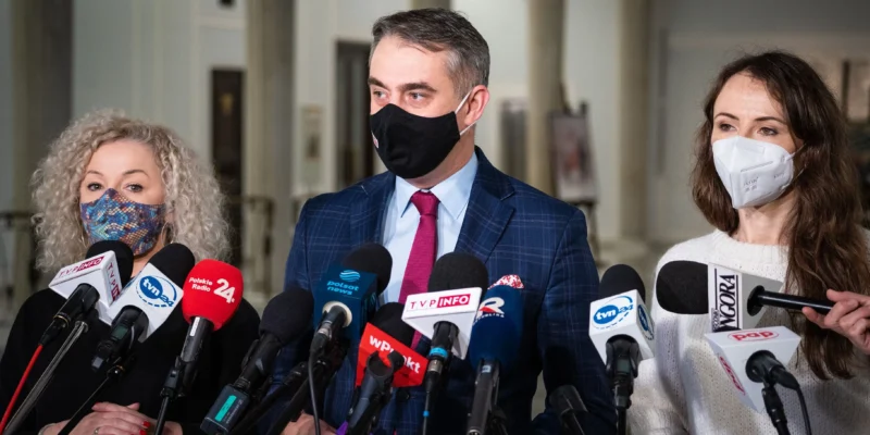 Dwie kobiety i jeden mężczyzna w maseczkach udzielają konferencji prasowej Lewicy, stojąc za zestawem mikrofonów.