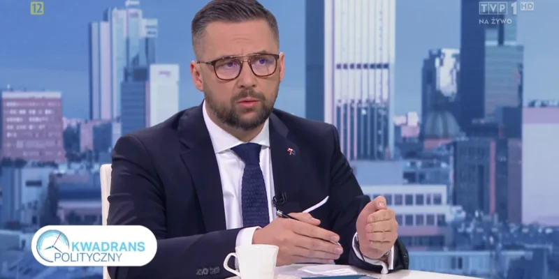 Mężczyzna w garniturze i okularach siedzi przy stole w studiu telewizyjnym podczas programu „Kwadrans Polityczny” emitowanego na żywo, z panoramą miasta w tle.