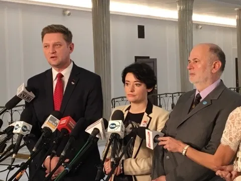 Trzy osoby w eleganckich strojach stoją przed wieloma mikrofonami podczas konferencji prasowej.