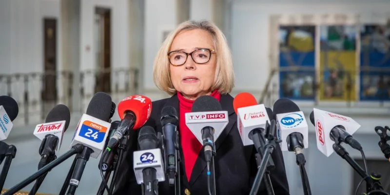 Kobieta w okularach, ubrana w czerwoną bluzkę i czarną marynarkę, przemawia w otoczeniu mikrofonów podczas konferencji prasowej.