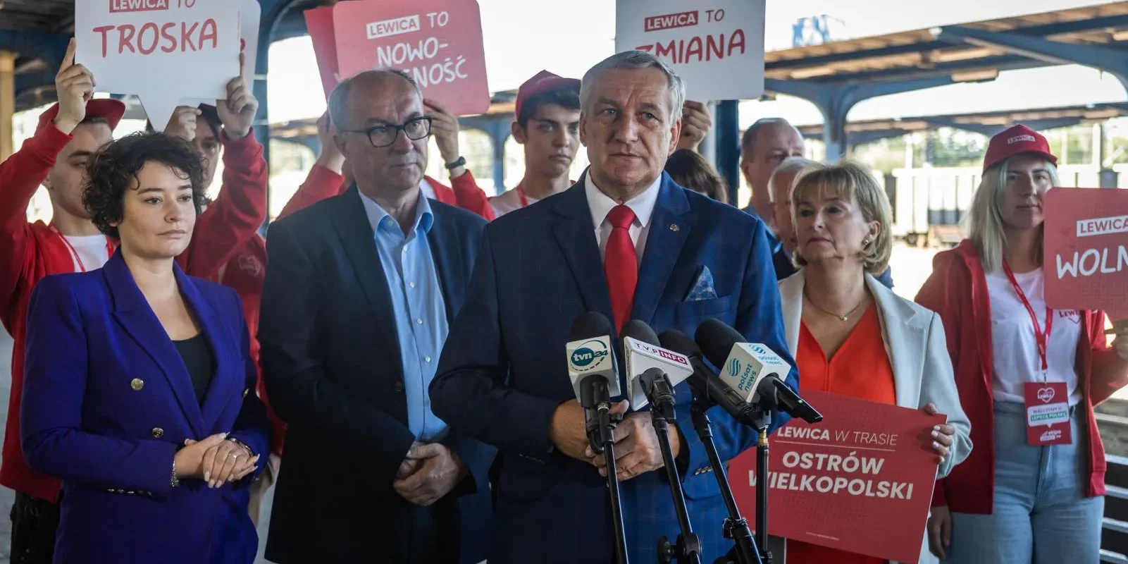 Mężczyzna w granatowym garniturze i czerwonym krawacie przemawia na konferencji prasowej, stojąc przed mikrofonami, otoczony przez zwolenników trzymających czerwone tablice z hasłami Lewica.