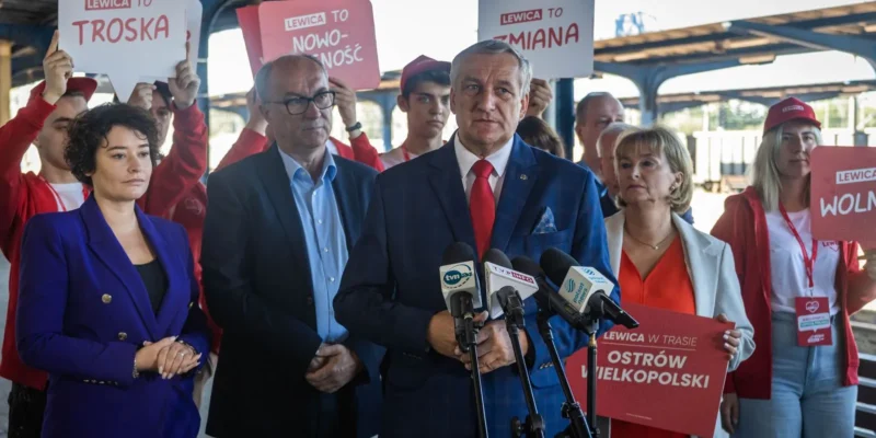 Mężczyzna w granatowym garniturze i czerwonym krawacie przemawia na konferencji prasowej, stojąc przed mikrofonami, otoczony przez zwolenników trzymających czerwone tablice z hasłami Lewica.