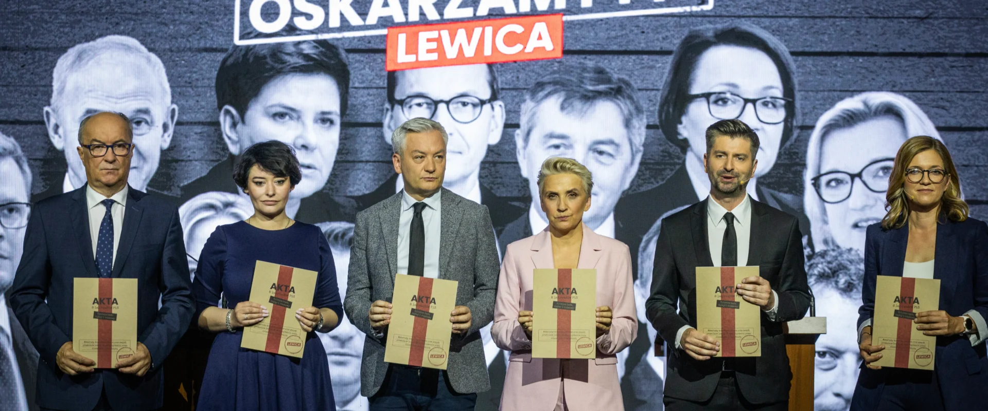 Szóstka polityków stoi na scenie w garniturach i eleganckich ubraniach, trzymając dokumenty podpisane „AKTA”, na tle czarno‑białego muralu z hasłem „OSKARŻAMY PIS LEWICA”.