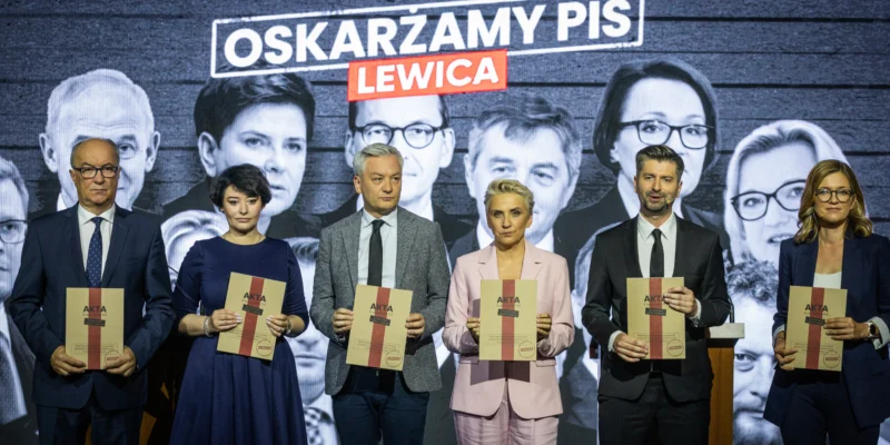 Szóstka polityków stoi na scenie w garniturach i eleganckich ubraniach, trzymając dokumenty podpisane „AKTA”, na tle czarno‑białego muralu z hasłem „OSKARŻAMY PIS LEWICA”.