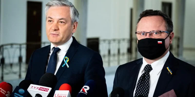 Dwaj mężczyźni w garniturach stoją przed mikrofonami podczas konferencji prasowej w jasnym wnętrzu.