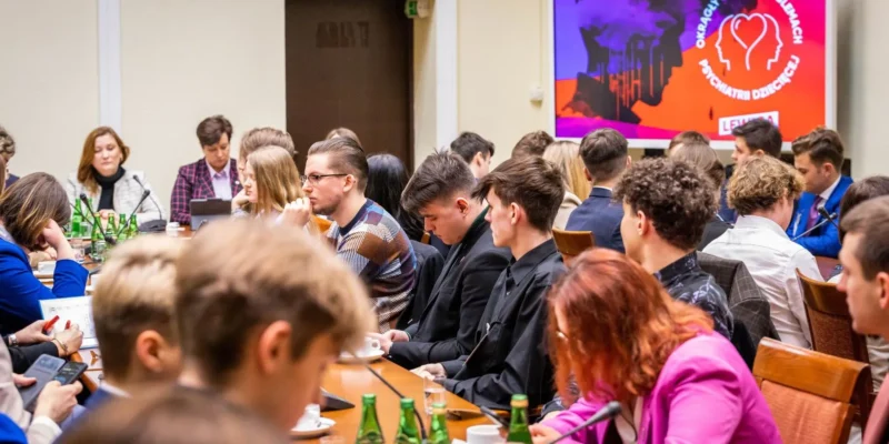 Zdjęcie przedstawia grupę młodych ludzi uczestniczących w dyskusji przy okrągłym stole, z wyświetlaczem w tle informującym o temacie spotkania.