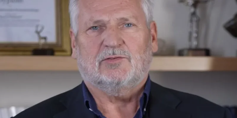 Mężczyzna o siwych włosach i brodzie, ubrany w granatowy garnitur z pinem w barwach flagi, siedzi przed półką z certyfikatami; w prawym górnym rogu widoczny jest logotyp Lewica Lista nr 3.