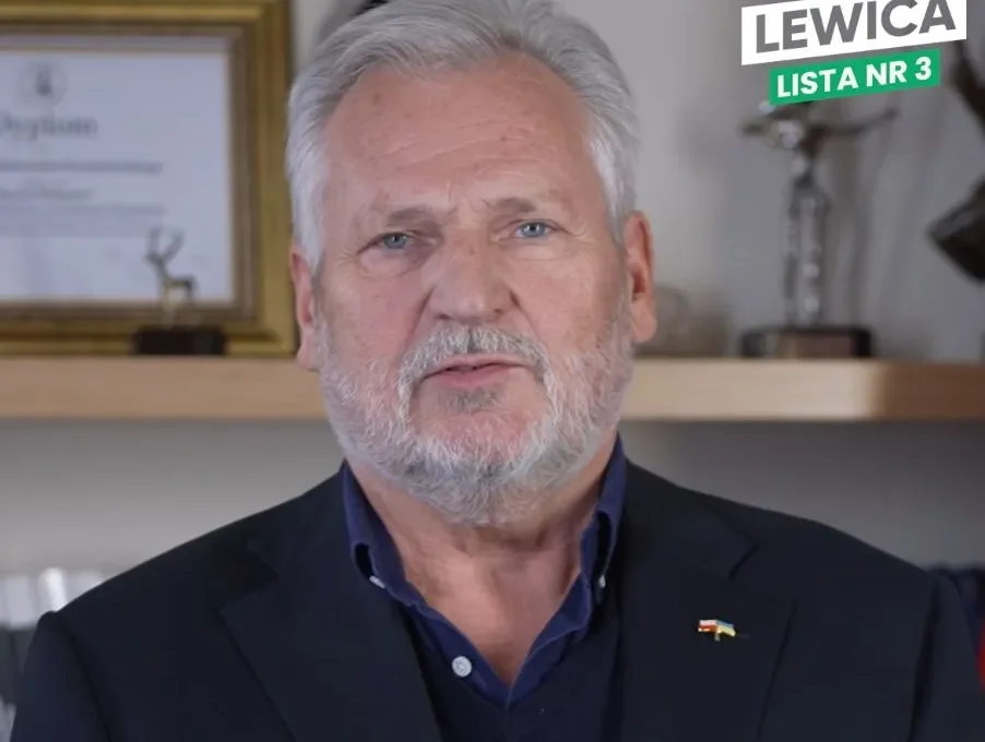 Mężczyzna o siwych włosach i brodzie, ubrany w granatowy garnitur z pinem w barwach flagi, siedzi przed półką z certyfikatami; w prawym górnym rogu widoczny jest logotyp Lewica Lista nr 3.