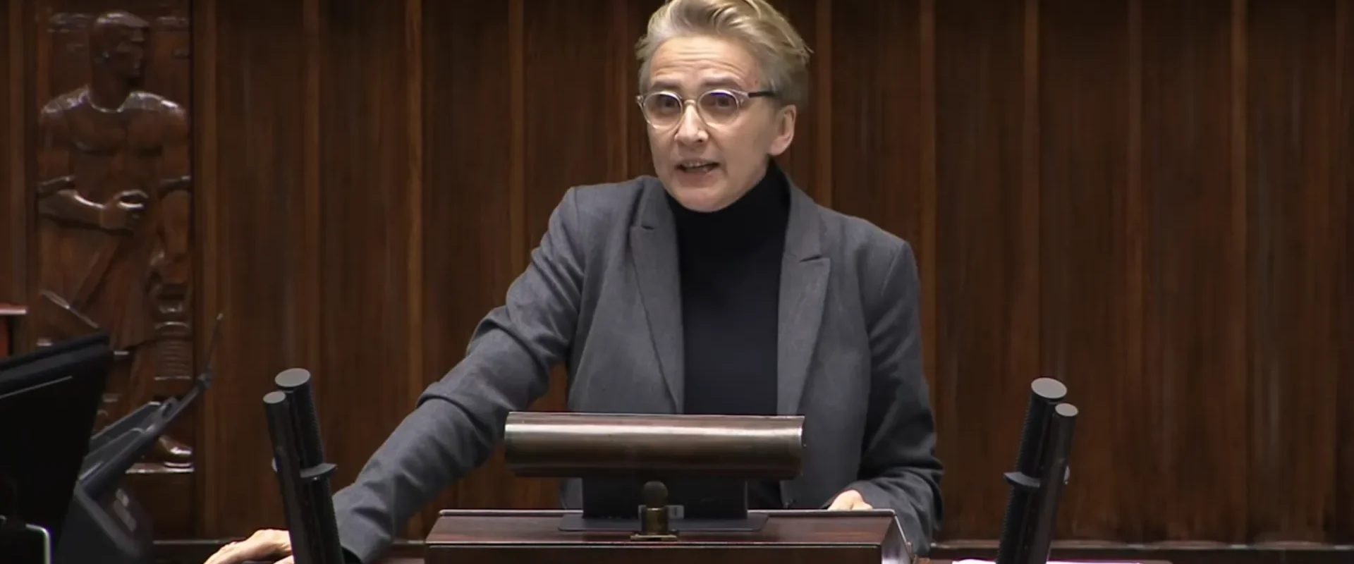 Na zdjęciu przedstawiona jest kobieta w okularach, występująca za mównicą w sali obrad, podczas gdy z tyłu widoczna jest drewniana ściana z rzeźbami.