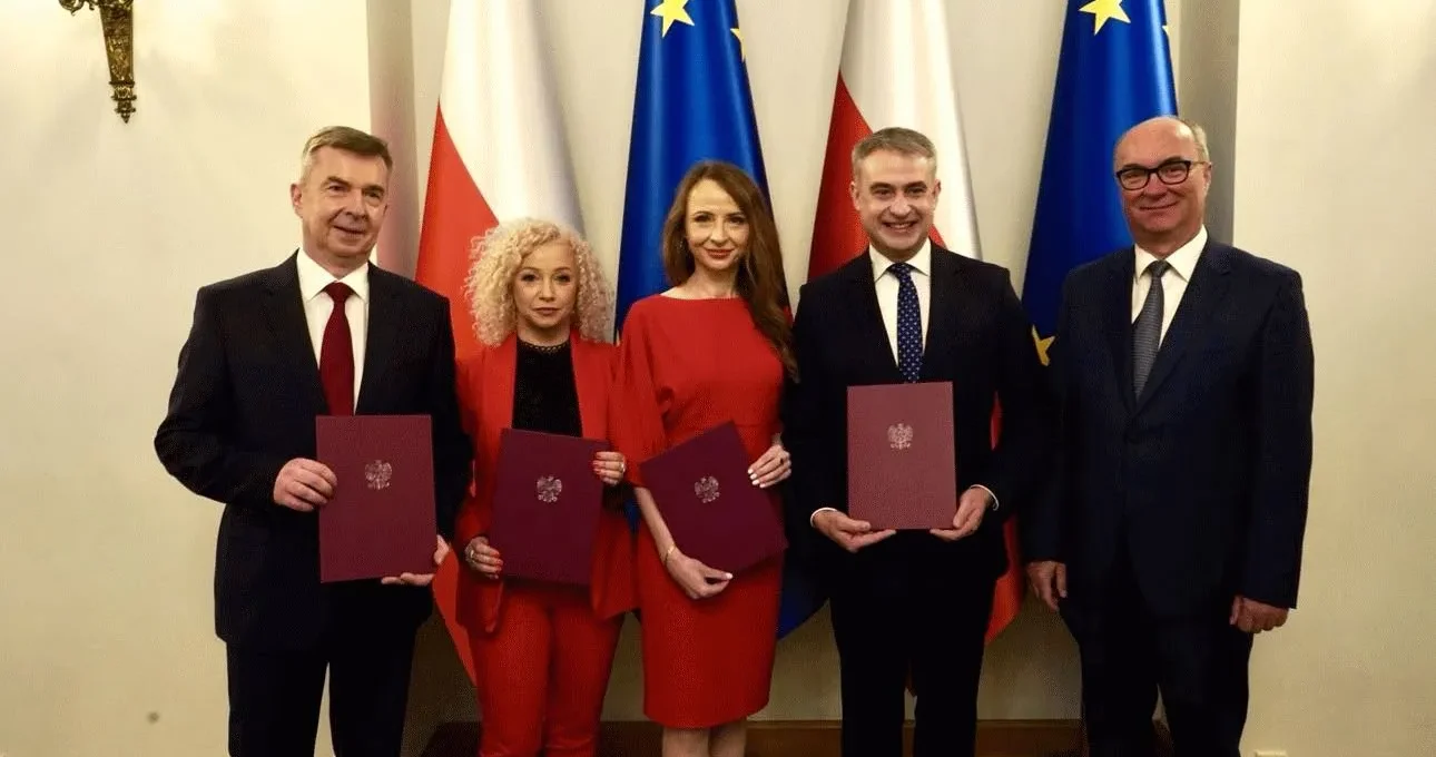Pięć osób w eleganckich garniturach i czerwonych strojach trzyma dokumenty podczas zaprzysiężenia rządu, stojąc na tle flag Polski i Unii Europejskiej.