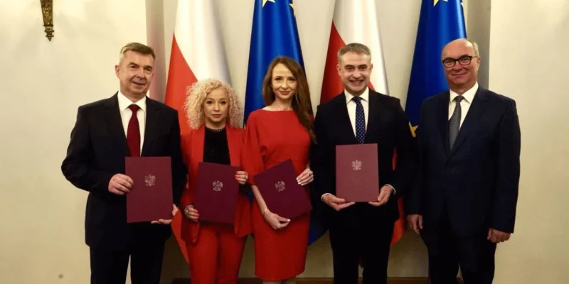 Pięć osób w eleganckich garniturach i czerwonych strojach trzyma dokumenty podczas zaprzysiężenia rządu, stojąc na tle flag Polski i Unii Europejskiej.