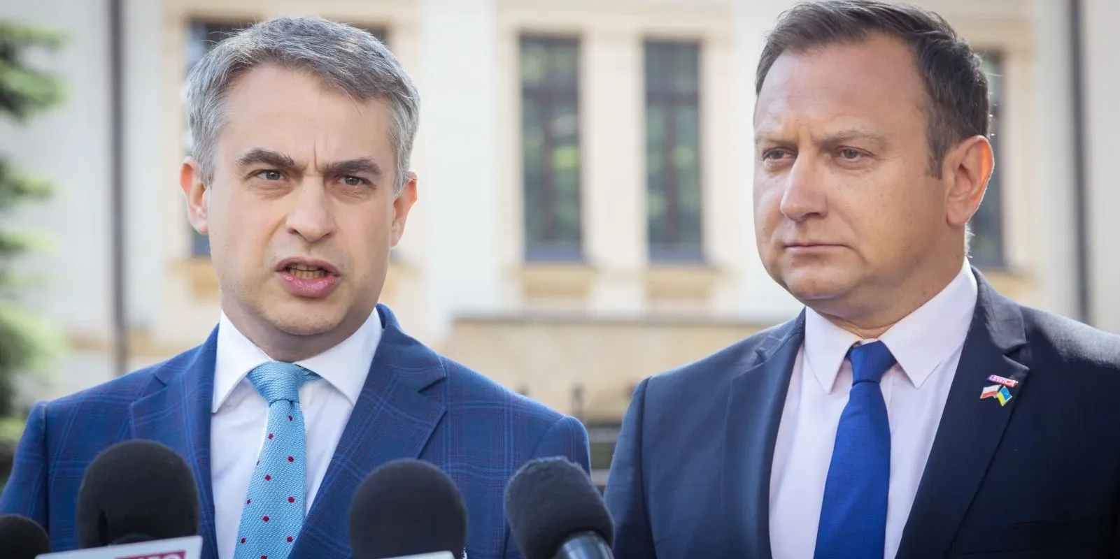 Dwóch mężczyzn w garniturach udziela konferencji prasowej, stojąc przed licznymi mikrofonami na tle budynku.