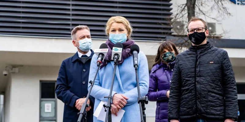 Grupa osób w maseczkach stoi przy mikrofonach podczas konferencji prasowej na zewnątrz, na tle budynku widoczna jest kobieta w jasnoniebieskim płaszczu.