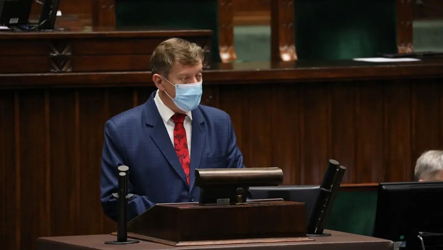 Mężczyzna w granatowym garniturze i niebieskiej maseczce stoi przy mównicy w sali obrad.