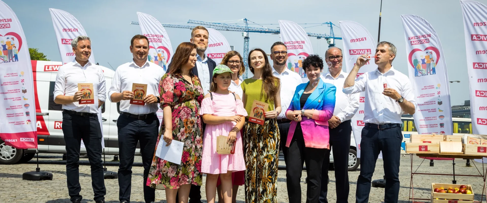 Grupa kilkunastu osób w białych koszulach i kolorowych ubraniach stoi na placu podczas wydarzenia „Wakacyjna trasa Lewicy – Bezpieczna Rodzina”, trzymając broszury i uśmiechając się wśród banerów i pojazdu z logo Lewicy w tle.