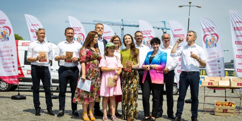 Grupa kilkunastu osób w białych koszulach i kolorowych ubraniach stoi na placu podczas wydarzenia „Wakacyjna trasa Lewicy – Bezpieczna Rodzina”, trzymając broszury i uśmiechając się wśród banerów i pojazdu z logo Lewicy w tle.