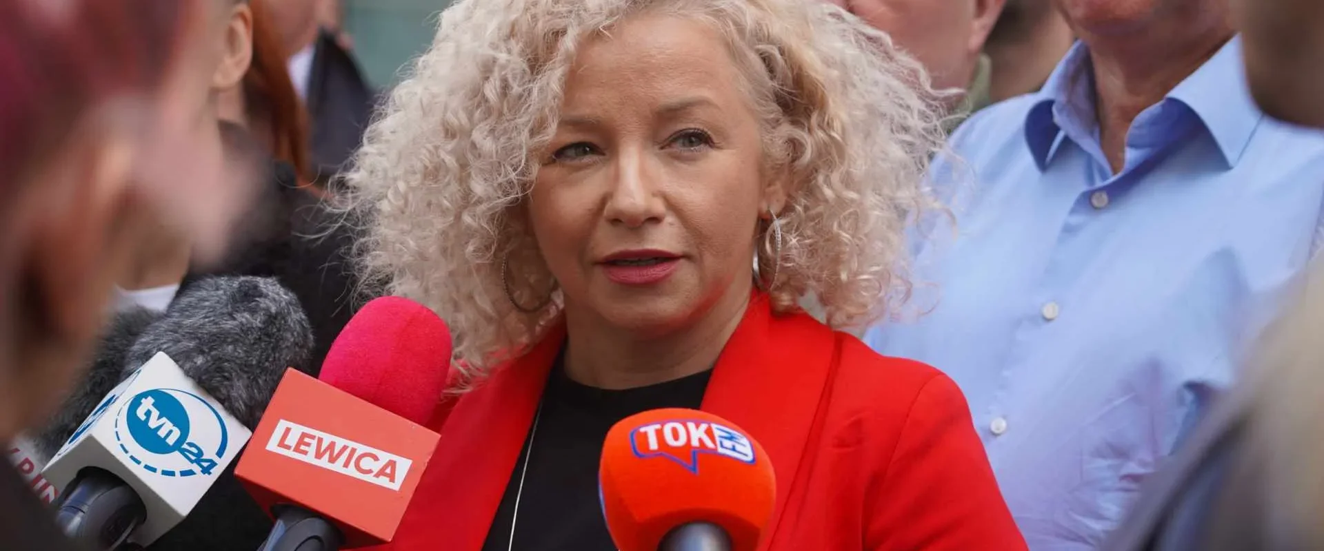 Kobieta o kręconych blond włosach w czerwonej marynarce udziela wywiadu przed kamerami podczas konferencji prasowej, otoczona dziennikarzami i mikrofonami z logotypami Lewicy.