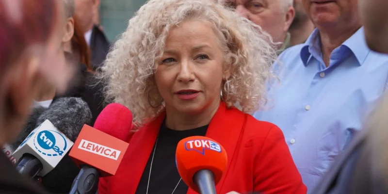 Kobieta o kręconych blond włosach w czerwonej marynarce udziela wywiadu przed kamerami podczas konferencji prasowej, otoczona dziennikarzami i mikrofonami z logotypami Lewicy.
