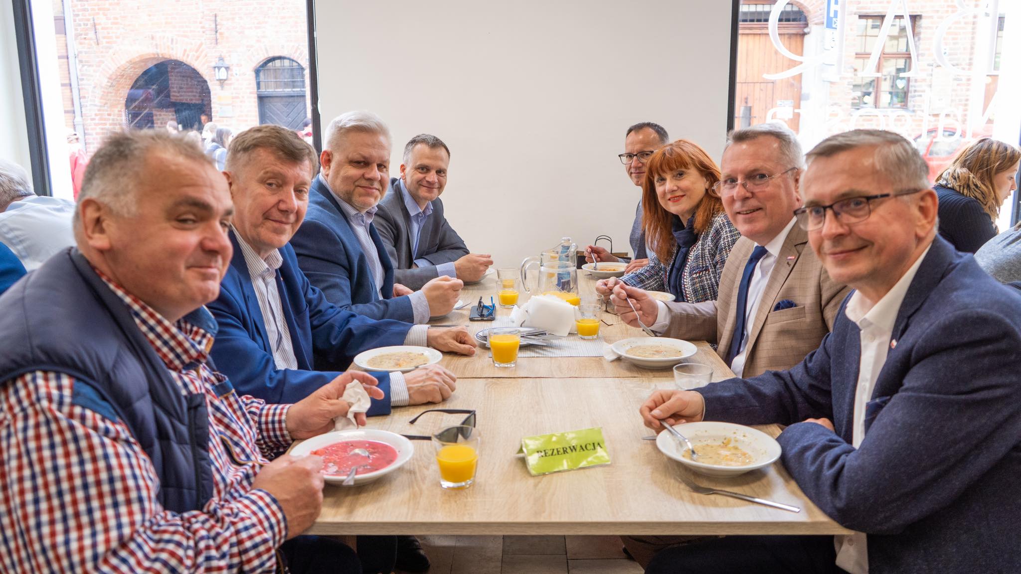 Zdjęcie przedstawia grupę ludzi siedzących przy stole w barze mlecznym, degustujących posiłki i rozmawiających ze sobą.