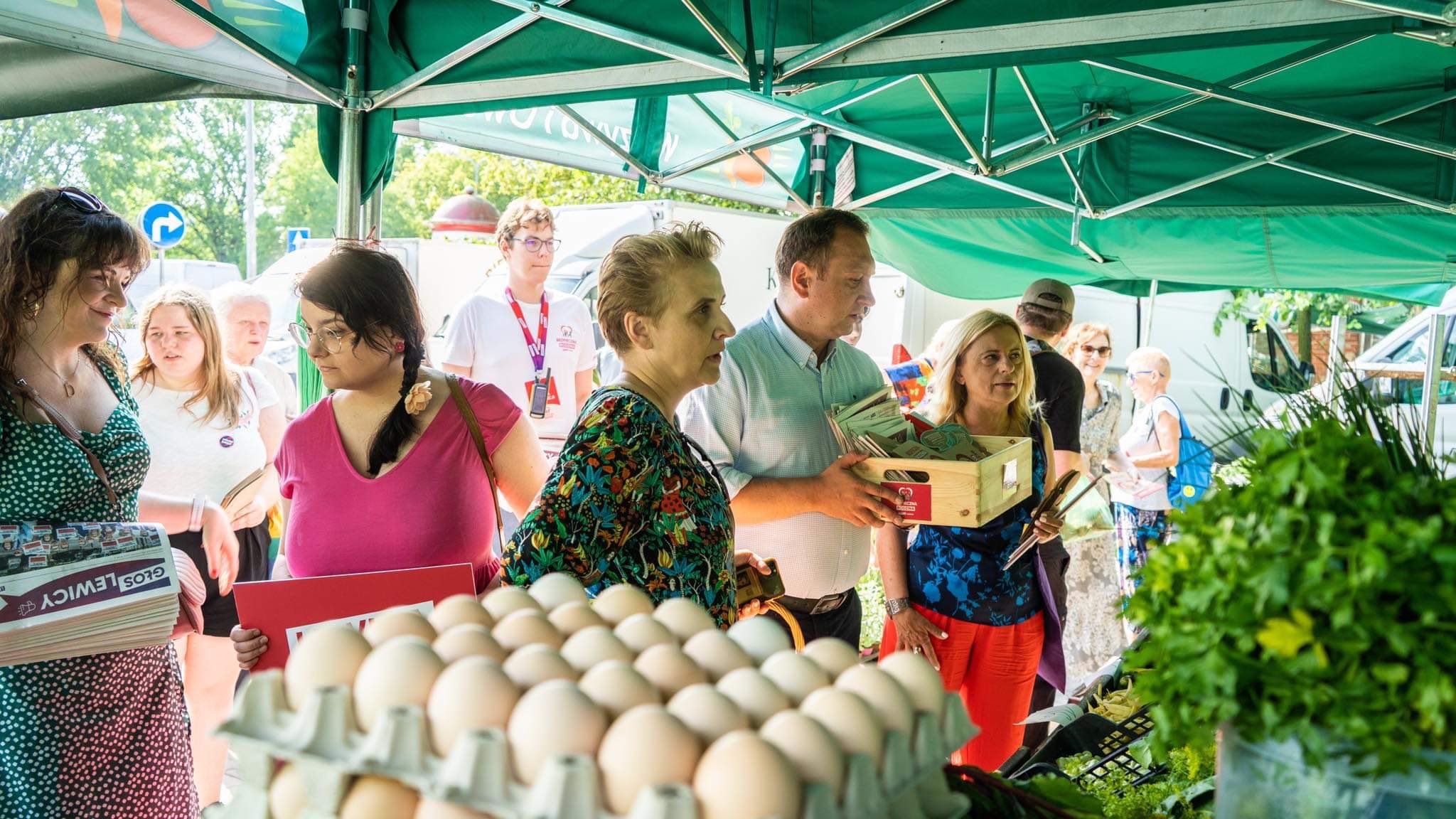 Na zdjęciu widać grupę ludzi kupujących produkty na bazarze, z jajkami na pierwszym planie oraz zielonymi warzywami w tle.