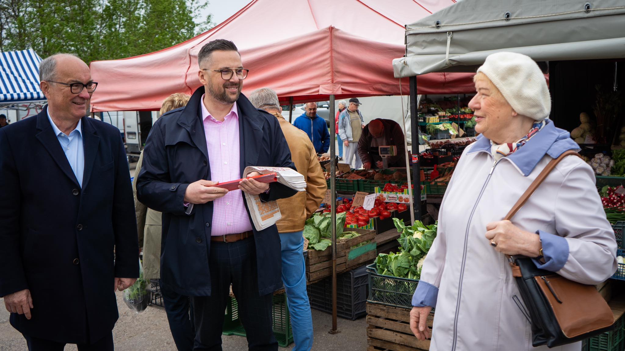 Na zdjęciu widać grupę osób rozmawiających na bazarze, w otoczeniu straganów z warzywami i owocami.